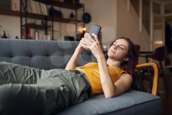 Young Woman Lying On Sofa At Home Looking At Mobile Phone Messages And Social Media