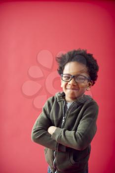 Portrait Of Boy Wearing Glasses With Folded Arms Against Red Studio Background Smiling At Camera