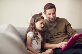 Father And Daughter Sitting On Sofa At Home Playing Together On Digital Tablet In Pink Case At Home