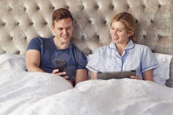 Couple At Home In Bed Self Isolating Using Digital Tablet And Mobile Phone During Covid 19 Lockdown