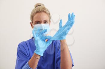 Studio Portrait Of Female Nurse Wearing Scrubs And PPE Face Mask Putting On Sterile Gloves