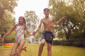 Children Wearing Swimming Costumes Having Fun In Garden Playing In Water From Garden Sprinkler