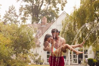 Father Carries Daughter Through Water From Garden Sprinkler Having Fun Wearing Swimming Costumes