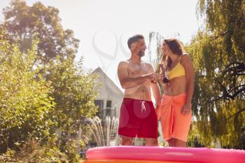 Couple Wearing Swimming Costumes Standing In Paddling Pool In Summer Garden At Home Drinking Beer
