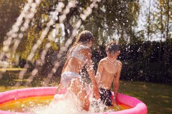 Children Wearing Swimming Costumes Having Fun In Garden Playing In Water From Garden Sprinkler