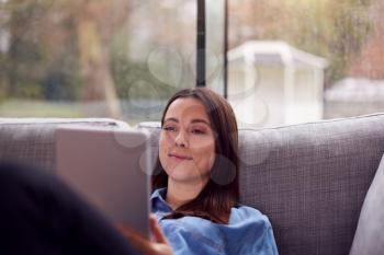 Smiling Young Woman At Home Lying On Sofa Looking At Digital Tablet