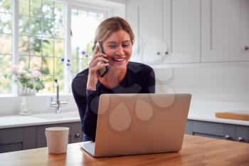 Woman In Kitchen With Mobile Phone Working From Home Using Laptop During Health Pandemic
