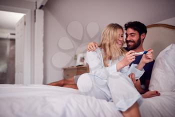 Excited Couple Wearing Pyjamas In Bedroom Celebrating Positive Pregnancy Test