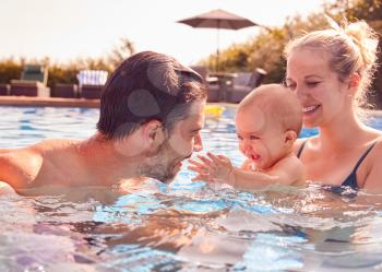 Family With Son And Baby Daughter Having Fun On Summer Vacation Splashing In Outdoor Swimming Pool