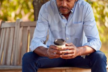 Close Up Of Mature Man Sitting On Park Bench Under Tree Holding Takeaway Coffee Cup