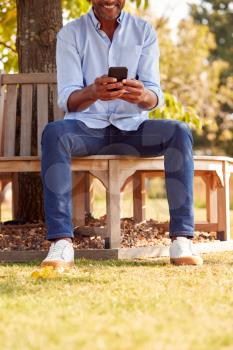 Close Up Of Man Sitting On Bench Under Tree In Summer Park Using Mobile Phone
