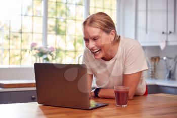 Mature Woman Wearing Fitness Clothing At Home Logging Activity From Smart Watch Onto Laptop