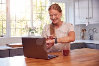 Mature Woman Wearing Fitness Clothing At Home Logging Activity From Smart Watch Onto Laptop