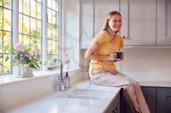 Mature Woman At Home With Hot Drink Sitting On Counter By Kitchen Window