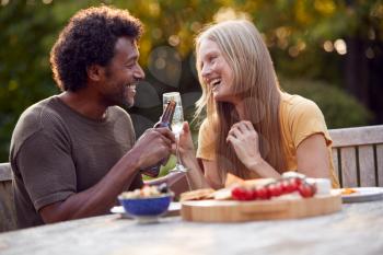 Mature Couple Celebrating With Beer And Champagne As They Sit At Table In Garden With Snacks