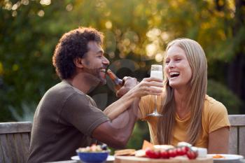 Mature Couple Link Arms Celebrating With Beer And Champagne Sitting At Table In Garden With Snacks