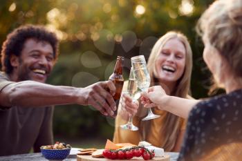 Group Of Friends Celebrating With Beer And Champagne As They Sit At Table In Garden With Snacks