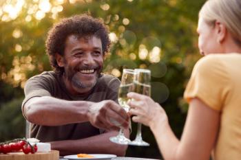 Mature Couple Celebrating With Champagne As They Sit At Table In Garden With Snacks