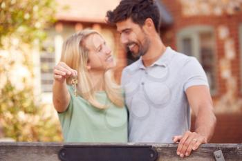 Portrait Of Excited Couple By Gate Holding House Keys Outside New Home In Countryside On Moving Day
