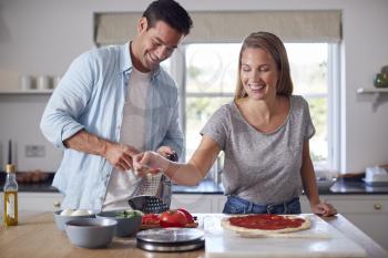 Woman Spreading Tomato Sauce On Base As Couple In Kitchen Home Prepare Homemade Pizzas Together