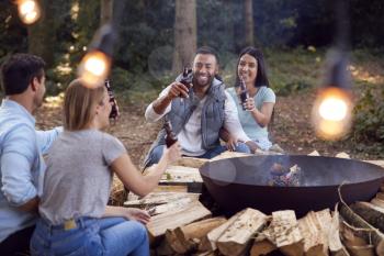 Group Of Friends Camping Sitting By Bonfire In Fire Bowl Celebrating And Drinking Beer Together