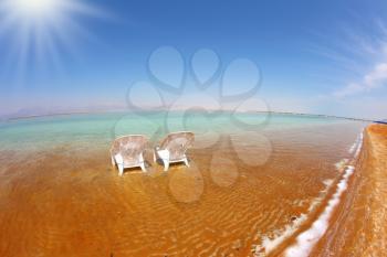 Two white beach chairs stood side by side in the clear water. The sun shines on the beach therapeutic