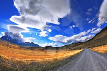 Gray dirt road in the Chile National Park Torres del Paine. Incredible shaped cloud formed by glaciers glisten in the sun. On the horizon are seen mountains with snow-capped peaks. Picture taken wit