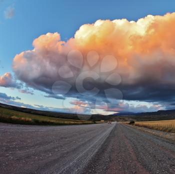 Huge pink - orange cloud, like a bowl of cream, lit the last rays of sunset. Gravel road through the Patagonian steppe