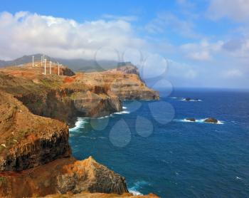 Over a cliff on the ocean breeze are the windmills. Eastern tip of the island of Madeira. Rocks steeply in the blue waters of the Atlantic Ocean. 