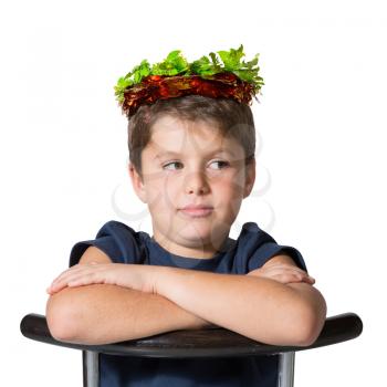 Boy sits astride a chair in carnival wearing a crown of shiny green and red leaf