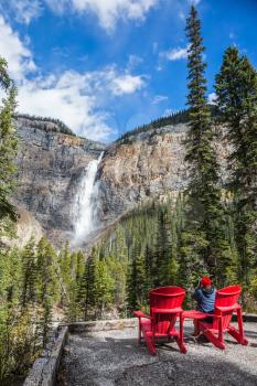 The woman photographs the famous waterfall. Two red deck chairs to relax in front of the waterfall Takakkaw. Autumn day in Yoho National Park in the Rocky Mountains 