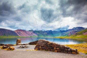The table and benches for country picnic.  Multi-colored mountains surround lake with ice-blue water