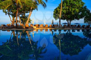 Sunset on the beach resort. An oceanfront pool surrounded by palm trees. Palm trees reflected in smooth water