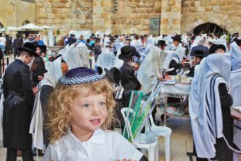 Adorable little boy with long blond curls and blue eyes in knitted skullcap. He stands at Western Wall of Temple. The Jewish holiday of Sukkot