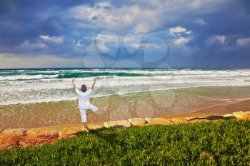 The beach in Tel Aviv. Storm cloud hanging over the sea, the woman in white performs asana Tree on one leg