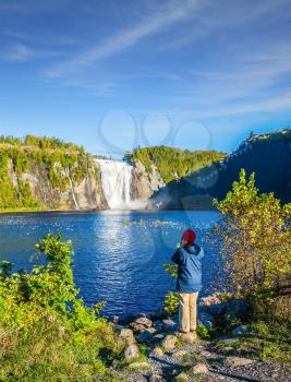 Woman in blue jacket with enthusiasm photographs waterfall Montmorency in Montmorency Falls Park. The concept of active and cultural tourism
