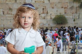 Cute little boy with long blond curls and blue eyes in knitted skullcap. He stands at the main Jewish shrine - Western Wall of Temple. The Jewish holiday of Sukkot