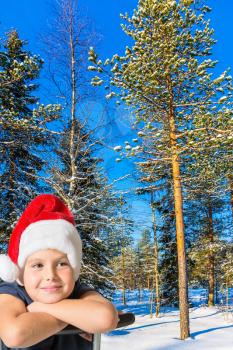Cold winter sunset. Soon Christmas. Charming boy in a red Santa Claus hat is smiling in a snow-covered winter forest