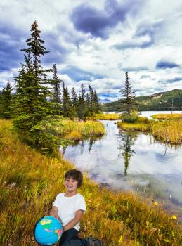 The concept of ecological and active tourism. Handsome boy with a globe in his hands laughs cheerfully in delight from clouds and lakes. Magnificent landscape in the Rocky Mountains