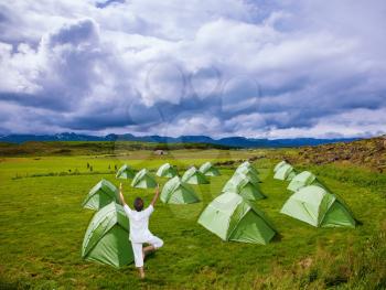 The woman in white clothes performs asana Tree. Campground athletes. Green tent on a grassy lawn