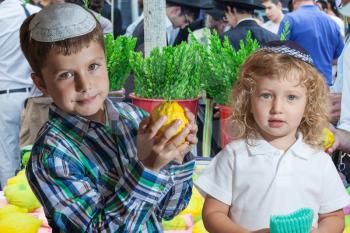 The Jewish holiday of Sukkot. Cute little boy with long blond hair in  knitted skullcap and  seven year old boy in white skullcap with etrog. They stand  on a pre-holiday market in Jerusalem. Ritual plants - myrtle prepared for sale

