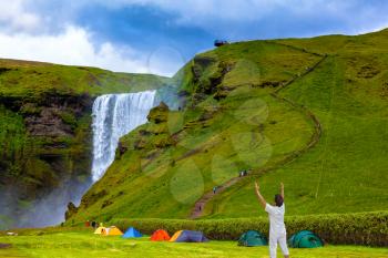 Grand waterfall Skogafoss. Near a waterfall put some colorful tourist tents. Elderly woman shocked by the beauty of the waterfall