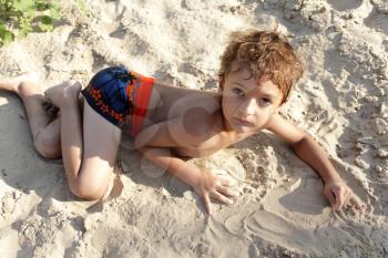 Portrait of happy little boy lying on the beach with his family in background while playing - Outdoors