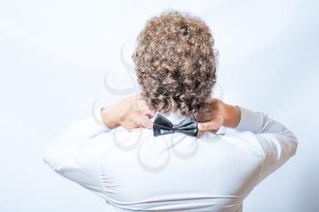 Bow tie on the back side. Strangeness or fun concept. Back view of an elegant young fashion man in tuxedo on gray background, toned image. Touch bow tie by hands.