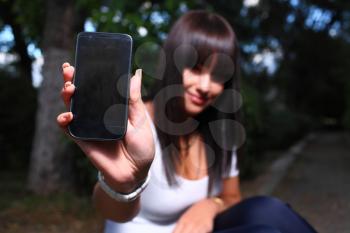 Smiling teenage girl shows cell phone, against green of summer park in the dark