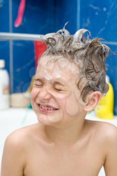 Smiling little boy front view. Kid covered with soap bubbles against a blue wall in bathroom. Closed eyes