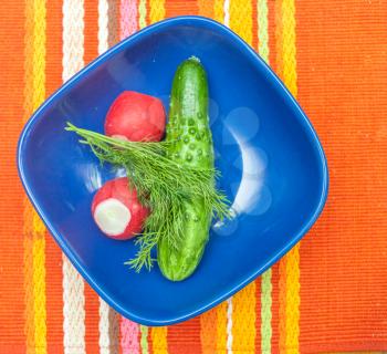 Radish, cucumber in blue bowl on red table cloth up view