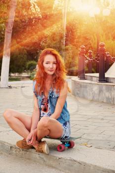 Red haired young women sitting on skateboard with her legs crossed backlit by sun.