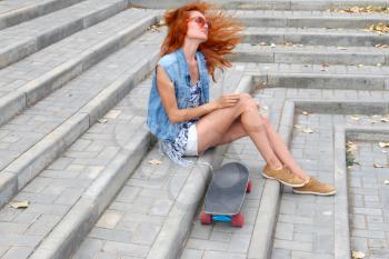Red haired girl sitting near her scateboard and her hair fly by wind