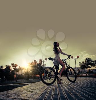 Biking evening. Attractive sporty slim girl sitting on bike on city alley looking back backlit, copyspace on sky.